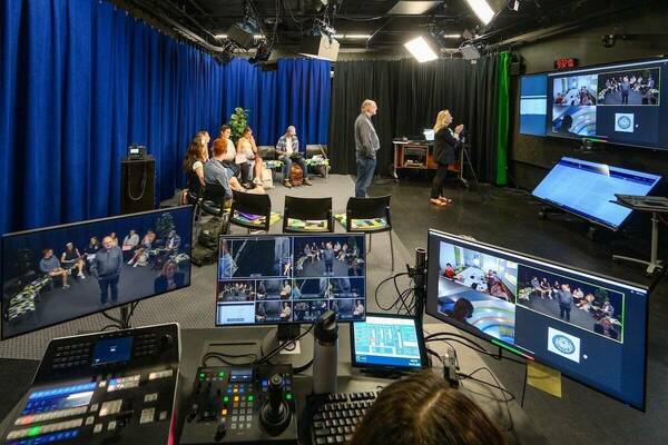 a media engineer at a station consisting of a variety of control panels and several monitors looks on as two professors teach a class with both in-person and remote students, the latter of whom are Zooming in via a large screen on the wall