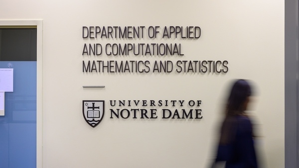 a blurred image of a person walking by a white wall with a large sign reading Department of Applied and Computational Mathematics and Statistics, University of Notre Dame