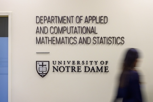 a blurred image of a person walking by a white wall with a large sign reading Department of Applied and Computational Mathematics and Statistics, University of Notre Dame