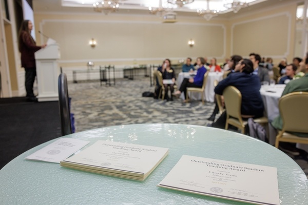 A photo of a ballroom, where in the foreground, there is a table with two stacks of Outstanding Graduate Student Teaching Award certificates. In the background, attendees look on as a speaker addresses them from a podium.