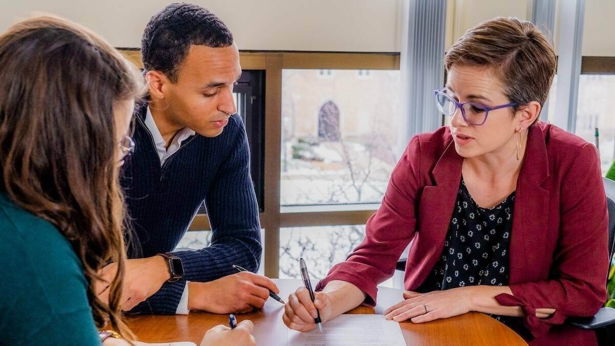 a woman sits at a table and makes notes on a piece of paper while two others look on