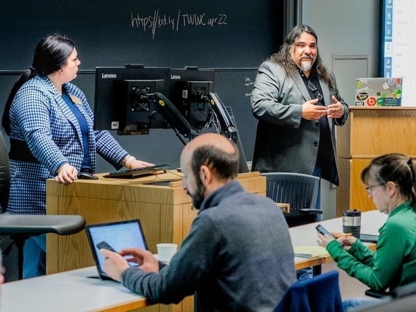 a man talks at a podium during a presentation in a seminar room while a woman looks on from a second podium and two members of the audience look something up on their phones