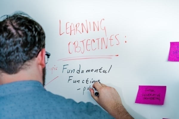 a man writes notes with a marker on a white board under the heading "Learning Objectives"