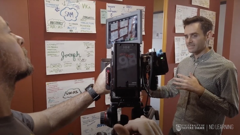 A photo of a man being filmed while talking next to a wall with pieces of paper pinned on it.