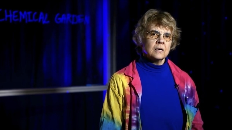 A professor stands in a darkened studio space delivering a lecture into a camera (unseen).