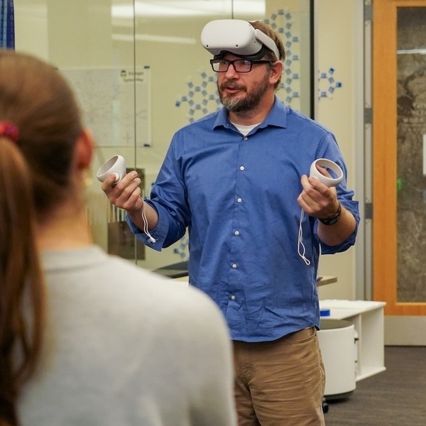 A man with a virtual reality headset resting atop his head and controllers in both of his hands explains to students how to use the technology.