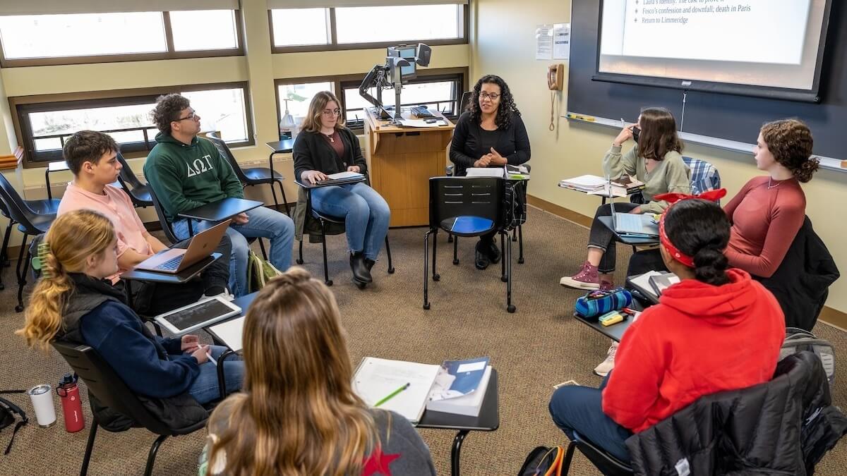 a professor and students sit together at desks arranged in a circle having a discussion