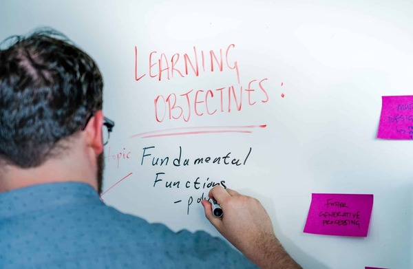 a man stands at a white board, on which he's written the words "Learning Objectives" in red