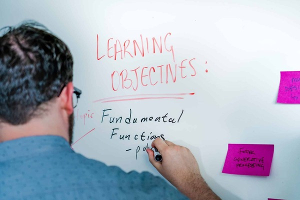 a man stands at a white board, on which he's written the words "Learning Objectives" in red