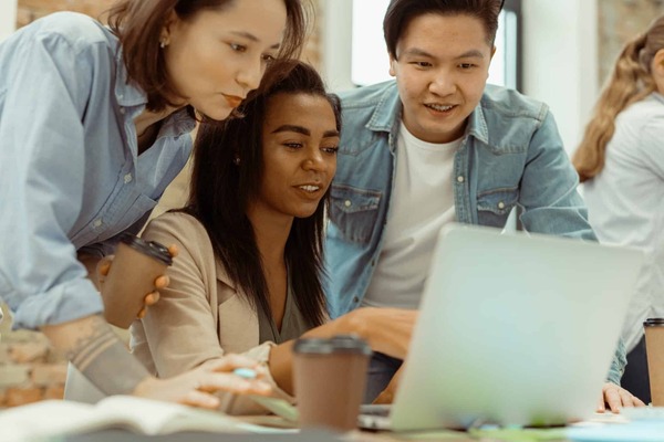 Three diverse students in casual clothes lean over a laptop and work together on a project.