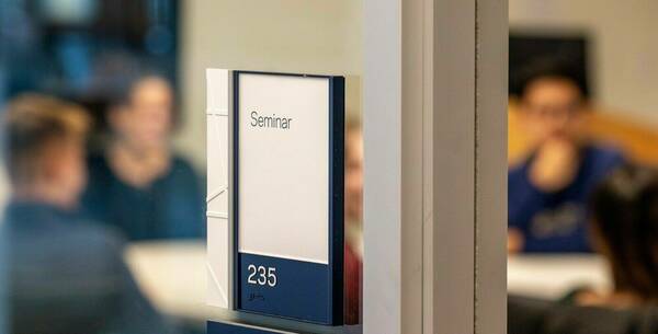 a photo of the exterior of a classroom, with a sign that says Seminar in the foreground and students through the windows in the background