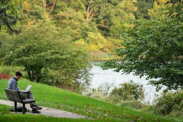 a student works on a laptop while sitting on a bench overlooking a lake