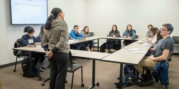 a professor stands at the front of a seminar-style classroom, where the tables are arranged in a rectangle, and listens to one of her students