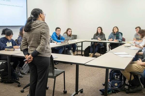 a professor stands at the front of a seminar-style classroom, where the tables are arranged in a rectangle, and listens to one of her students