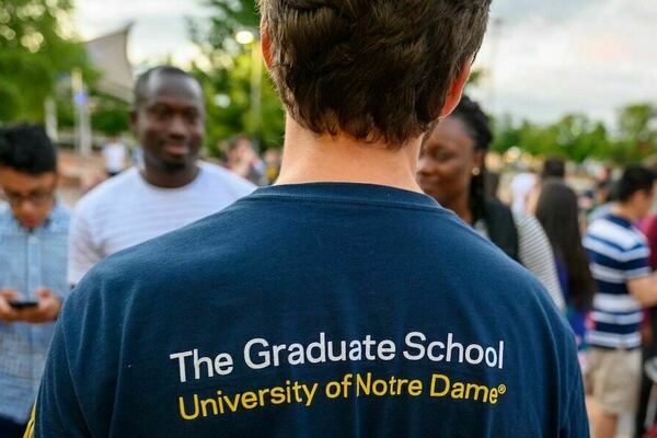 a student in a blue University of Notre Dame Graduate School t-shirt talks with other students outdoors at Graduate School Orientation