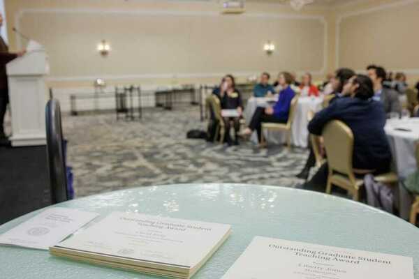 A photo of a ballroom, where in the foreground, there is a table with two stacks of Outstanding Graduate Student Teaching Award certificates. In the background, attendees look on as a speaker addresses them from a podium.