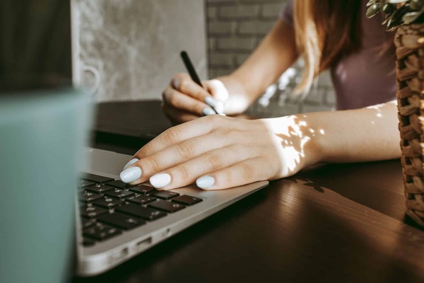 Close up of a woman's left hand resting on a laptop keyboard while her right hand holds a pen in the background.