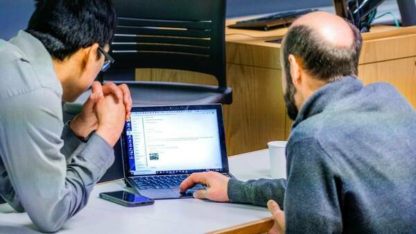 a man works on a laptop while another man leans in to look at the screen