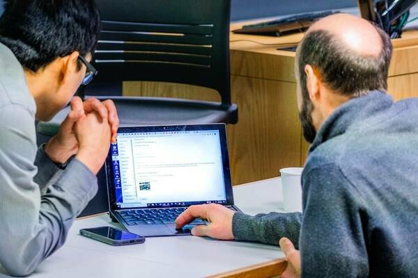 a man works on a laptop while another man leans in to look at the screen