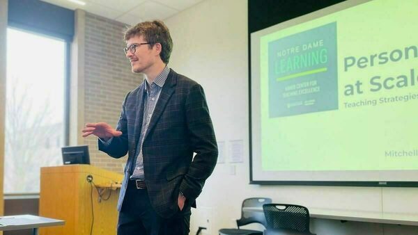 Dressed in a dark sports coat, Mitch Olsen speaks at the front of a seminar room. Visible on the screen on the wall behind him is part of the title slide for the workshop, "Personalization at Scale: Teaching Strategies for Large Class Sections."