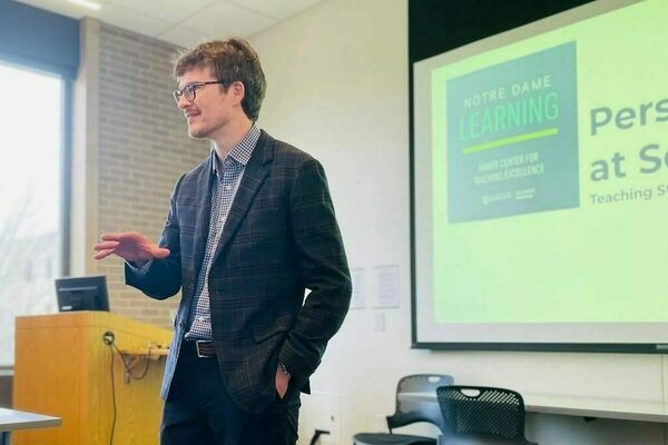 Dressed in a dark sports coat, Mitch Olsen speaks at the front of a seminar room. Visible on the screen on the wall behind him is part of the title slide for the workshop, "Personalization at Scale: Teaching Strategies for Large Class Sections."