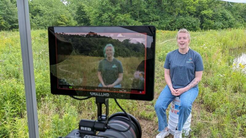 A person wearing a gray t-shirt and jeans sits on a bucket in a wetlands area and smiles at the camera.  A monitor displays their image.