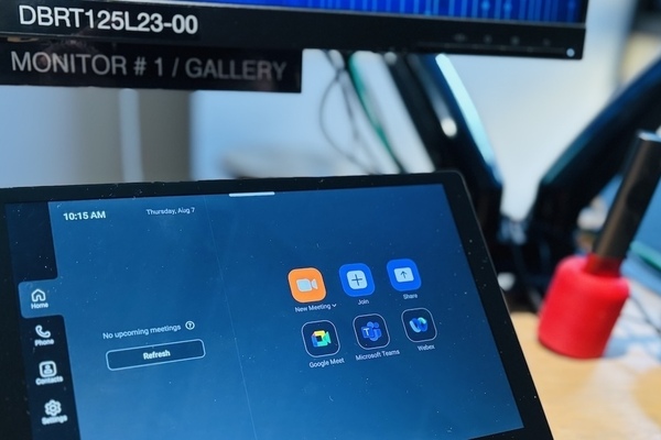 A Zoom Rooms touchscreen control panel displays video conferencing options, including New Meeting, Join, Share, Google Meet, Microsoft Teams, and Webex. A monitor labeled "DBRT125L23-00 MONITOR #1/GALLERY" is visible in the background.