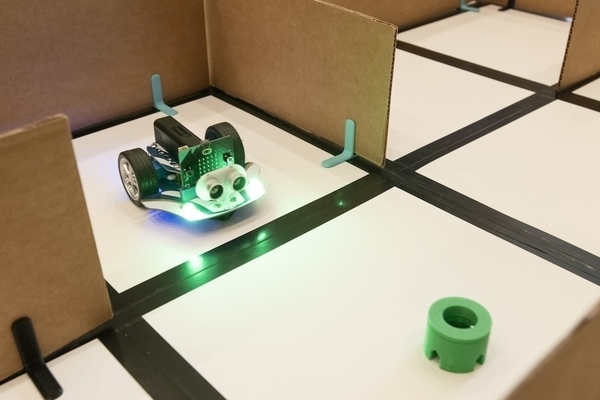 A small robot with glowing green lights navigates a cardboard maze toward a green plastic cylinder.