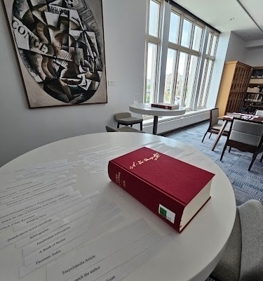 A red hardcover volume rests on a white circular table. White paper strips with research terms like "Encyclopedia Article" and "Bibliography" are scattered around it. The modern study room has large windows and an abstract painting on the wall.