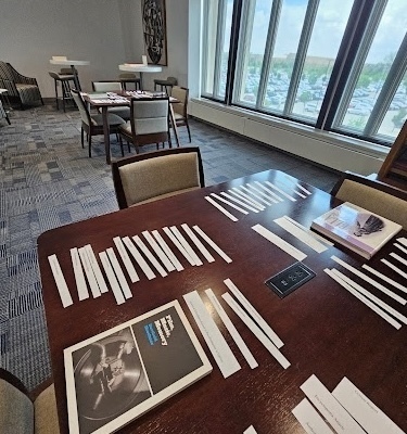 A large dark wooden table is covered with white paper strips and a book with a vinyl record on the cover. The modern room, furnished with tables and chairs, features large windows overlooking a parking lot and trees.