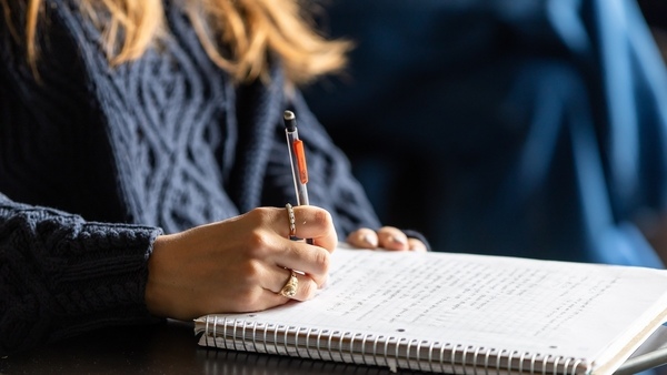 A closeup of a student's right hand writing in a spiral notebook. They have two rings on their fingers and are wearing a dark blue cable-knit sweater.