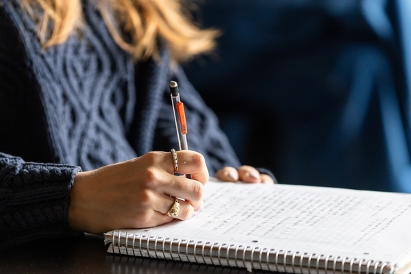 A closeup of a student's right hand writing in a spiral notebook. They have two rings on their fingers and are wearing a dark blue cable-knit sweater.