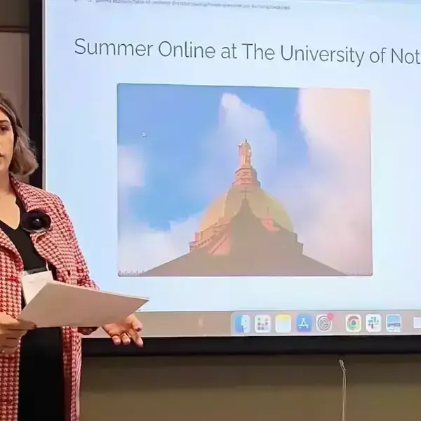 A woman in a red houndstooth jacket speaks, holding papers. The screen behind her shows Notre Dame's Golden Dome and a bulleted list of text describing the University's Summer Online program.