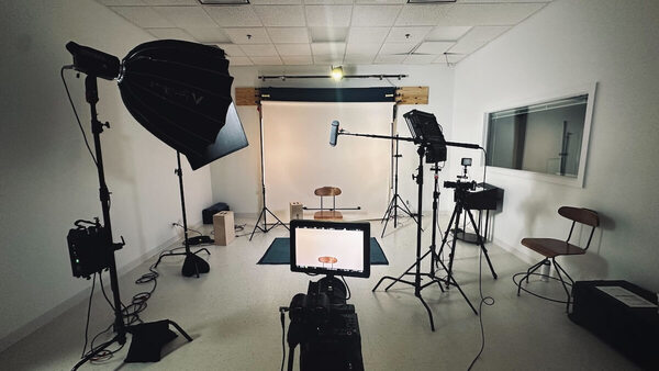 A video recording studio with a backdrop screen, lights on stands, and a camera pointed at a stool in front of the screen. The ceiling, walls, and floor are white, and the stool is visible in frame on the camera's display.