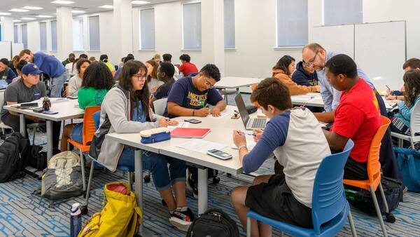 A large number of students work in small groups at tables in a bright classroom, using laptops, calculators, and pencil and paper. An instructor leans over to assist two at the nearest table.