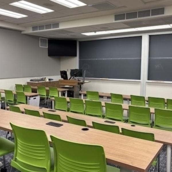 A view looking toward the front of an empty classroom that has green seats arranged at rectangular tables. Two monitors hang near the ceiling on the left, and small whiteboards hang from the ends of the tables.