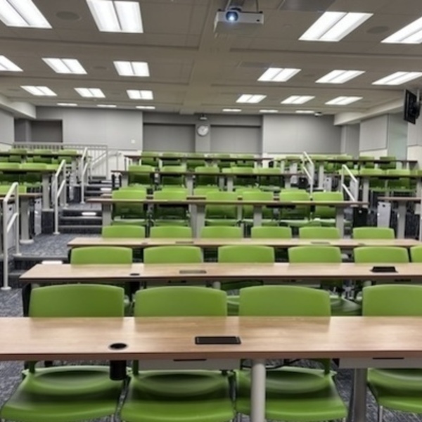 A view looking toward the back of an empty, tiered classroom with green seats broken into three columns at small rectangular tables. The columns form two aisles from the front to the back of the room, and whiteboards hang at the end of each table.