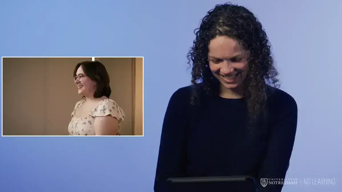 A screenshot from a video where a smiling professor is looking down at a tablet; she's watching a video of a student talking, which appears in an inset frame on the left side of the screen.