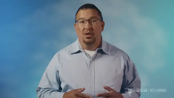 A man looks directly at the viewer in a still from a video, speaking and gesturing with his hands against a blue and teal background.