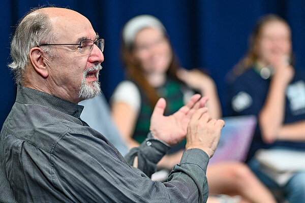 A professor gestures to a class while teaching. Two students are blurred in the background.