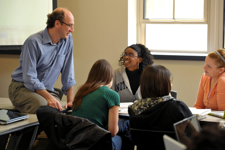 a professor talks with students seated at a table in class