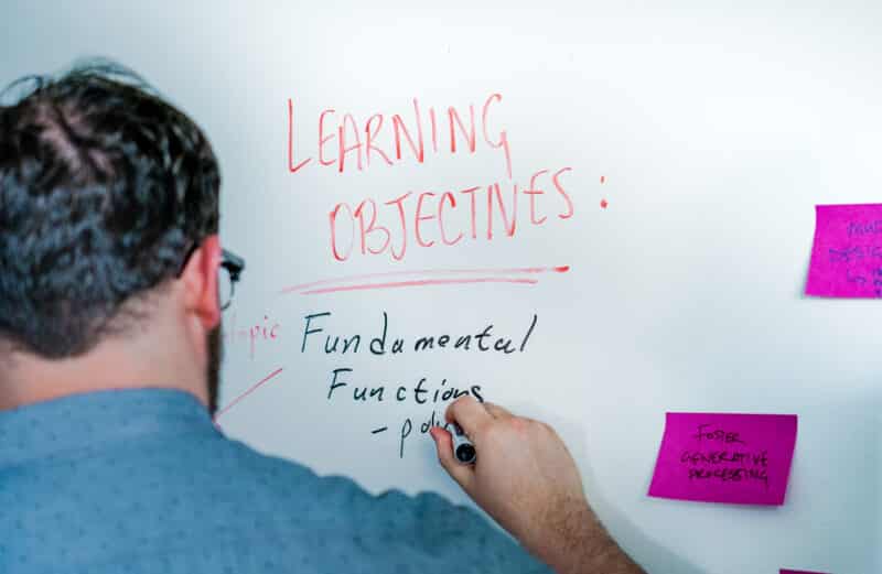 a man stands at a white board, on which he's written the words "Learning Objectives" in red