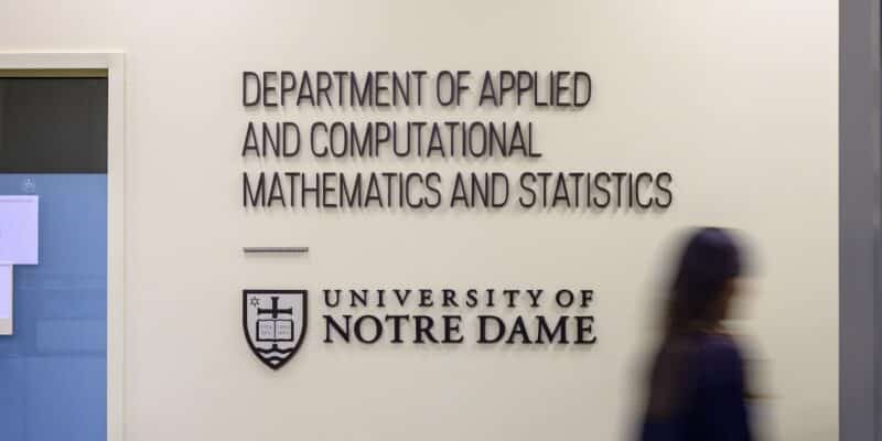 a blurred image of a person walking by a white wall with a large sign reading Department of Applied and Computational Mathematics and Statistics, University of Notre Dame