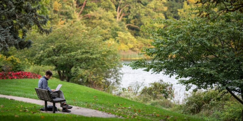 a student works on a laptop while sitting on a bench overlooking a lake