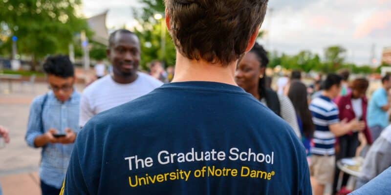 a student in a blue University of Notre Dame Graduate School t-shirt talks with other students outdoors at Graduate School Orientation