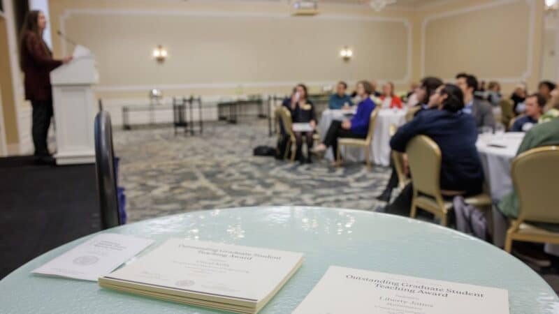 A photo of a ballroom, where in the foreground, there is a table with two stacks of Outstanding Graduate Student Teaching Award certificates. In the background, attendees look on as a speaker addresses them from a podium.