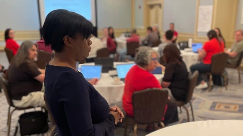 Veronica Womack stands next to a table and listens as an NDITA participant makes a comment during her interactive keynote session.